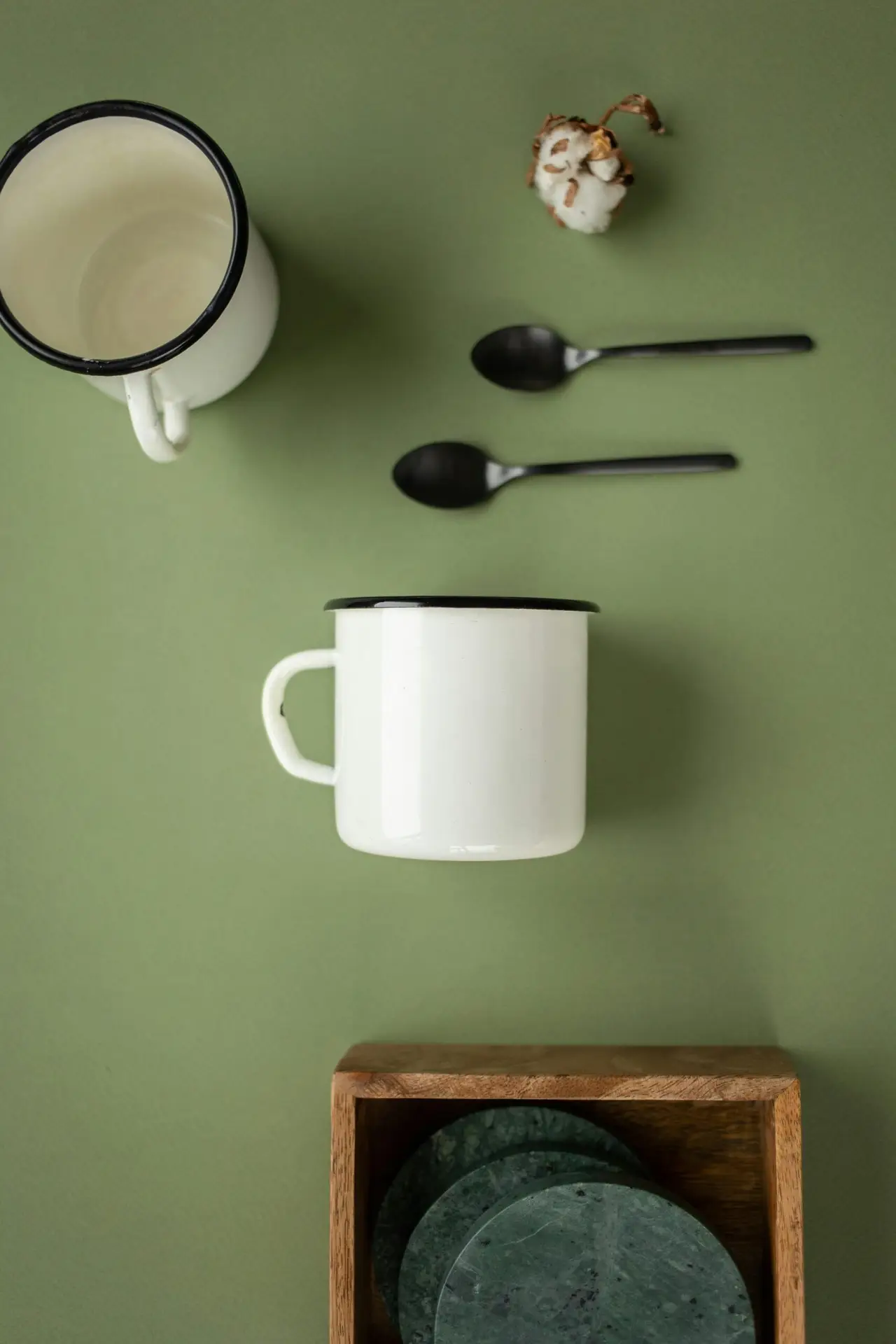 Top-view composition of cups, teaspoons, cotton flower, and wooden box on a green background.